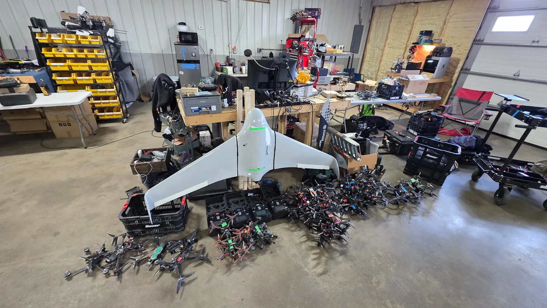 A fixed-wing aircraft and a fleet of FPV quads staged across a workshop floor.